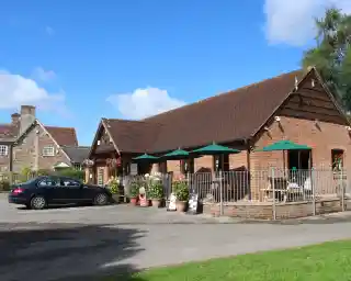 A charming brick building with a sloped roof features outdoor seating with green umbrellas. Colorful flower pots line the patio, and a black car is parked nearby. The scene is set against a blue sky with a few clouds.