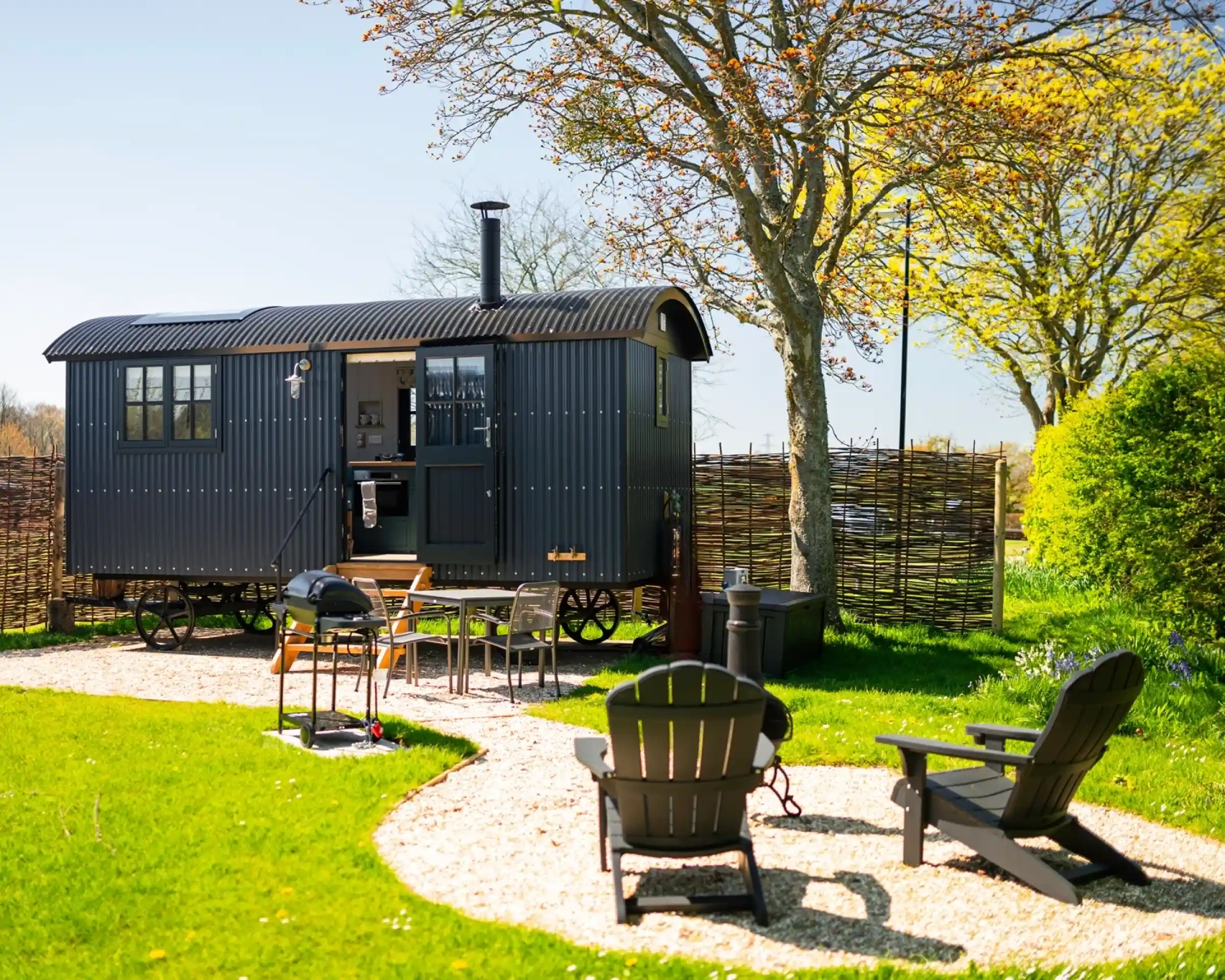 A charming black hut on wheels sits in a lush green area, surrounded by trees. In front, there are two wooden chairs and a small table, with a gravel path leading up to the hut. A warm sun shines down on the serene outdoor setting.