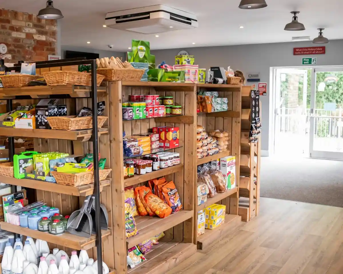Wooden shelves filled with various grocery items, including snacks, canned goods, and household products. The bright interior features exposed brick walls and a view of the outdoors through large glass doors.