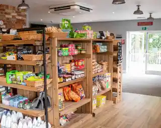 Wooden shelves filled with various grocery items, including snacks, canned goods, and household products. The bright interior features exposed brick walls and a view of the outdoors through large glass doors.