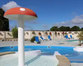 A vibrant swimming pool area featuring a large, red and white mushroom-shaped water feature surrounded by blue lounge chairs. The backdrop includes a wall with hanging flower baskets under a bright blue sky.