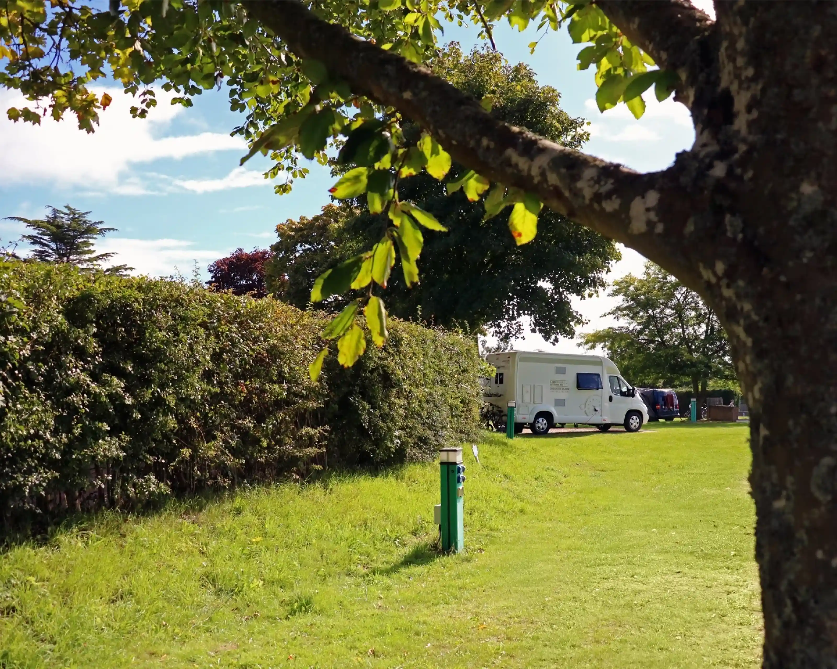 A grassy area with a tree in the foreground, a hedge in the background, and a white camper van parked nearby. Blue sky with some clouds is visible.