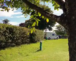 A grassy area with a tree in the foreground, a hedge in the background, and a white camper van parked nearby. Blue sky with some clouds is visible.