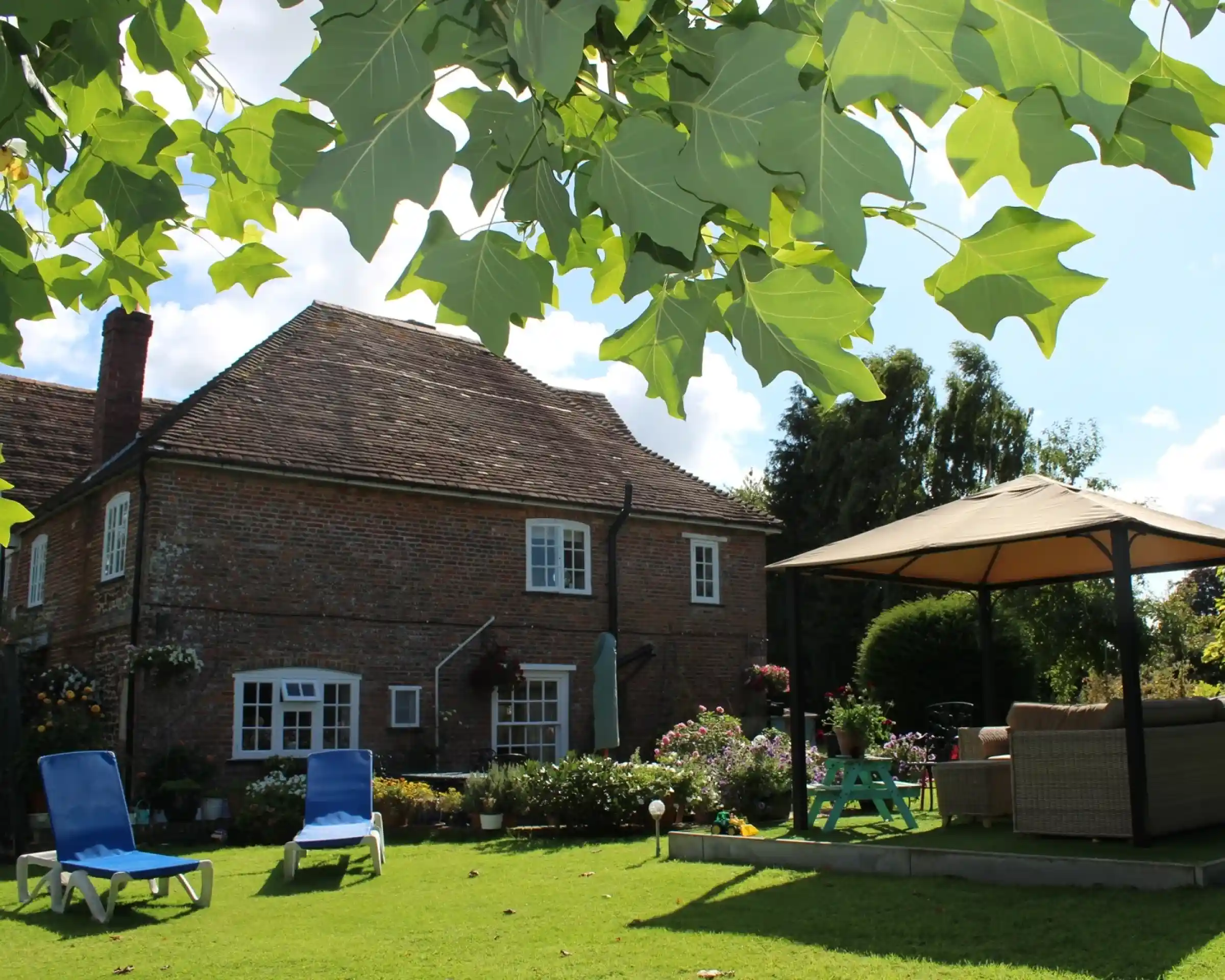 A charming brick house with multiple windows is surrounded by colorful flowers and greenery. In the foreground, two blue lounge chairs sit on a well-manicured lawn, alongside a shaded gazebo with outdoor furniture. Lush trees frame the scene under a bright blue sky.
