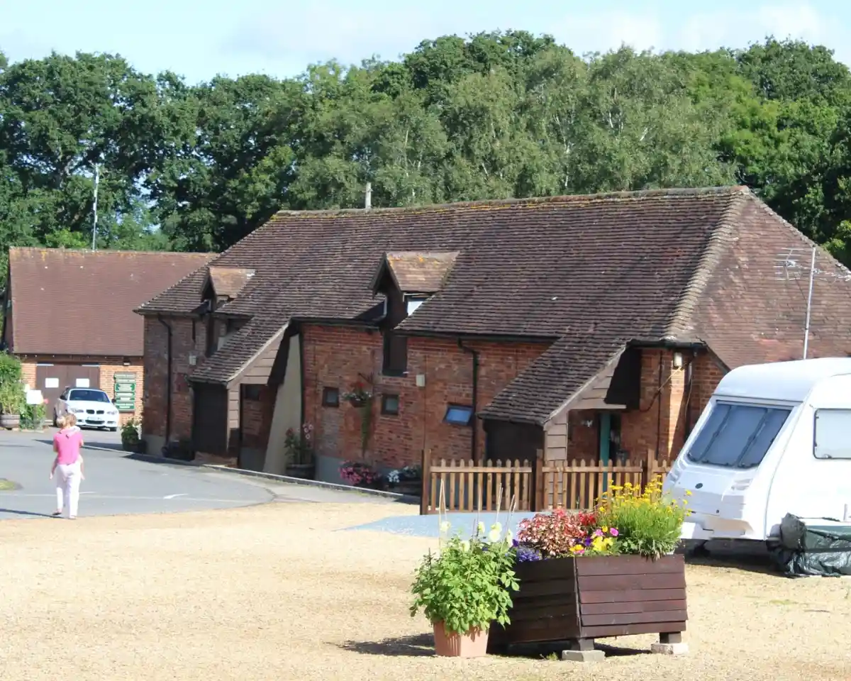 A rustic brick building with a tiled roof, surrounded by greenery and gravel pathways. A parked caravan is nearby, and a person walks toward the entrance. Colorful flower pots add charm to the scene.