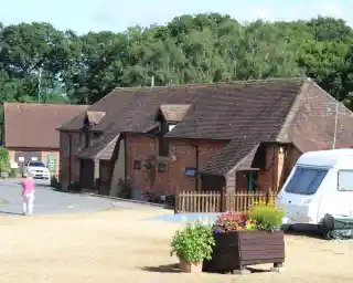 A rustic brick building with a tiled roof, surrounded by greenery and gravel pathways. A parked caravan is nearby, and a person walks toward the entrance. Colorful flower pots add charm to the scene.