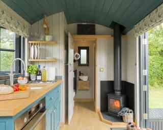 A cozy, modern kitchen interior featuring a blue and wooden color scheme. There is a small stove, a sink, and an oven, along with dishes neatly arranged. A wood-burning stove adds warmth, and a window shows a green outdoor view.