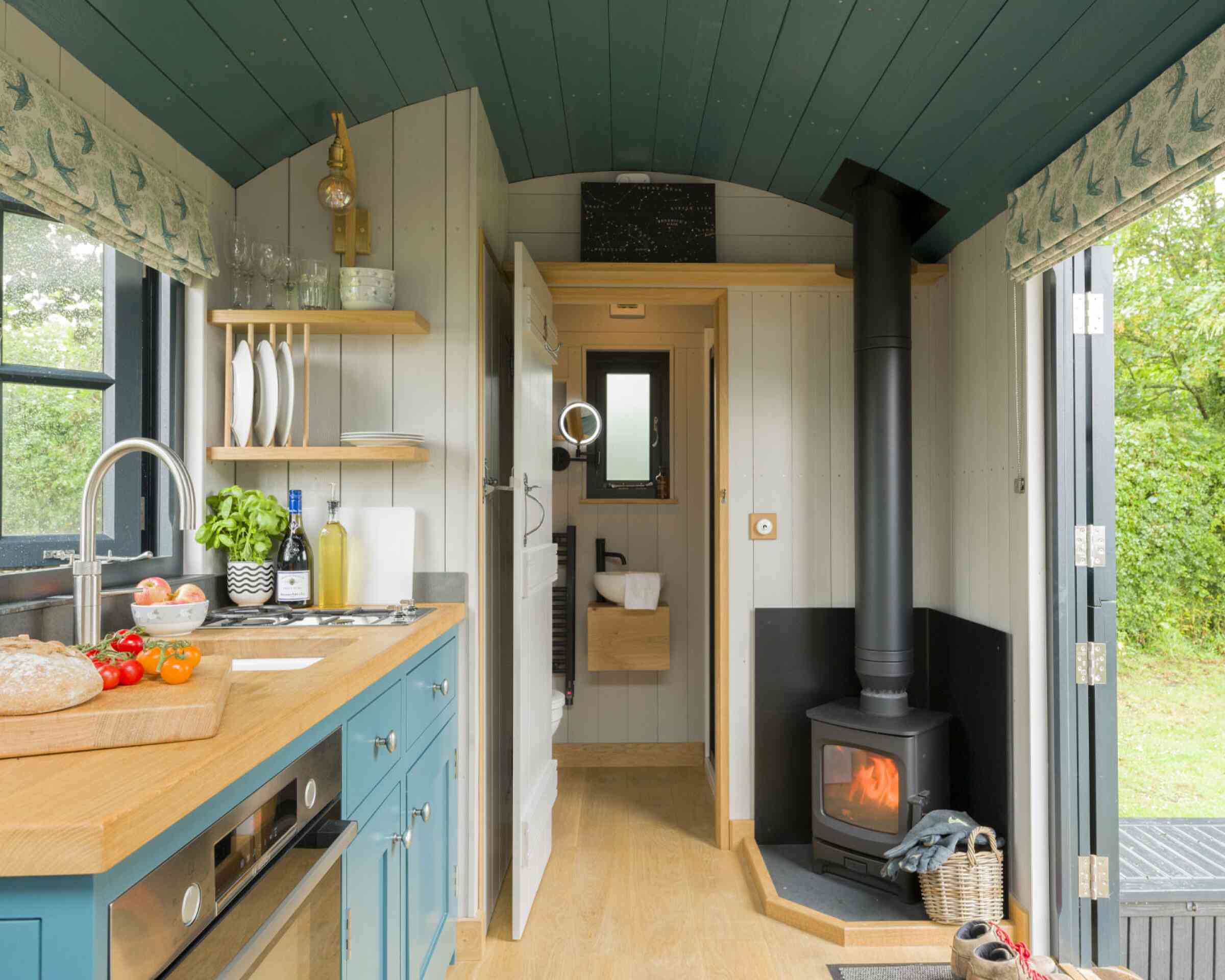 A cozy, modern kitchen interior featuring a blue and wooden color scheme. There is a small stove, a sink, and an oven, along with dishes neatly arranged. A wood-burning stove adds warmth, and a window shows a green outdoor view.