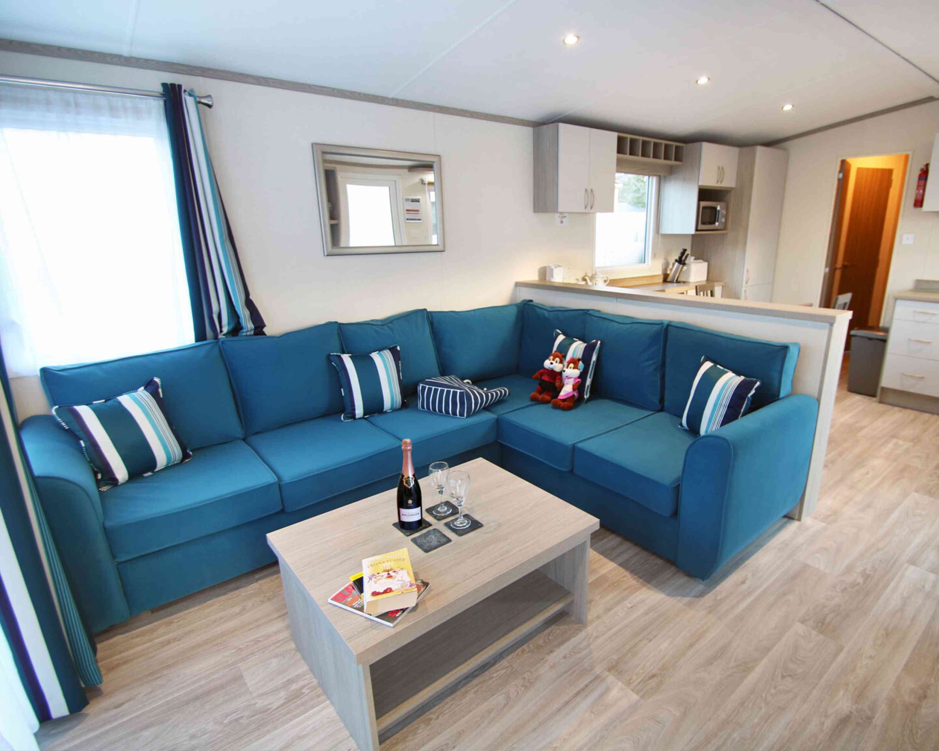 A cozy living area featuring a blue sectional sofa with decorative pillows, a light wooden coffee table with champagne and glasses, and a kitchen area visible in the background. Bright natural light enters through a window, enhancing the inviting atmosphere.