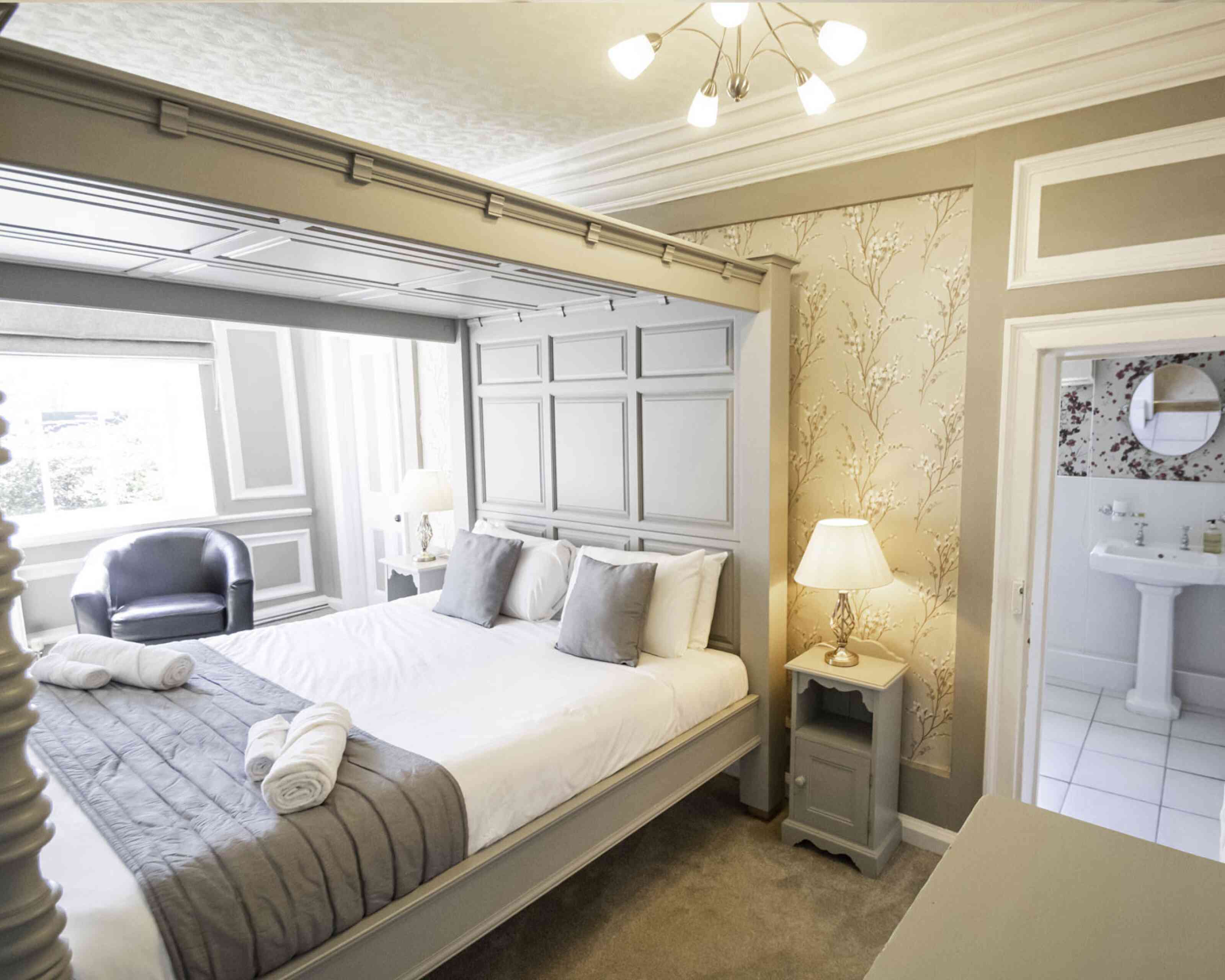 A stylish bedroom featuring a large gray bed with white linens and gray cushions. There’s a cozy armchair in the corner, a bedside lamp on a small table, and soft, neutral-toned decor with floral wallpaper. A bathroom is visible through an open door.