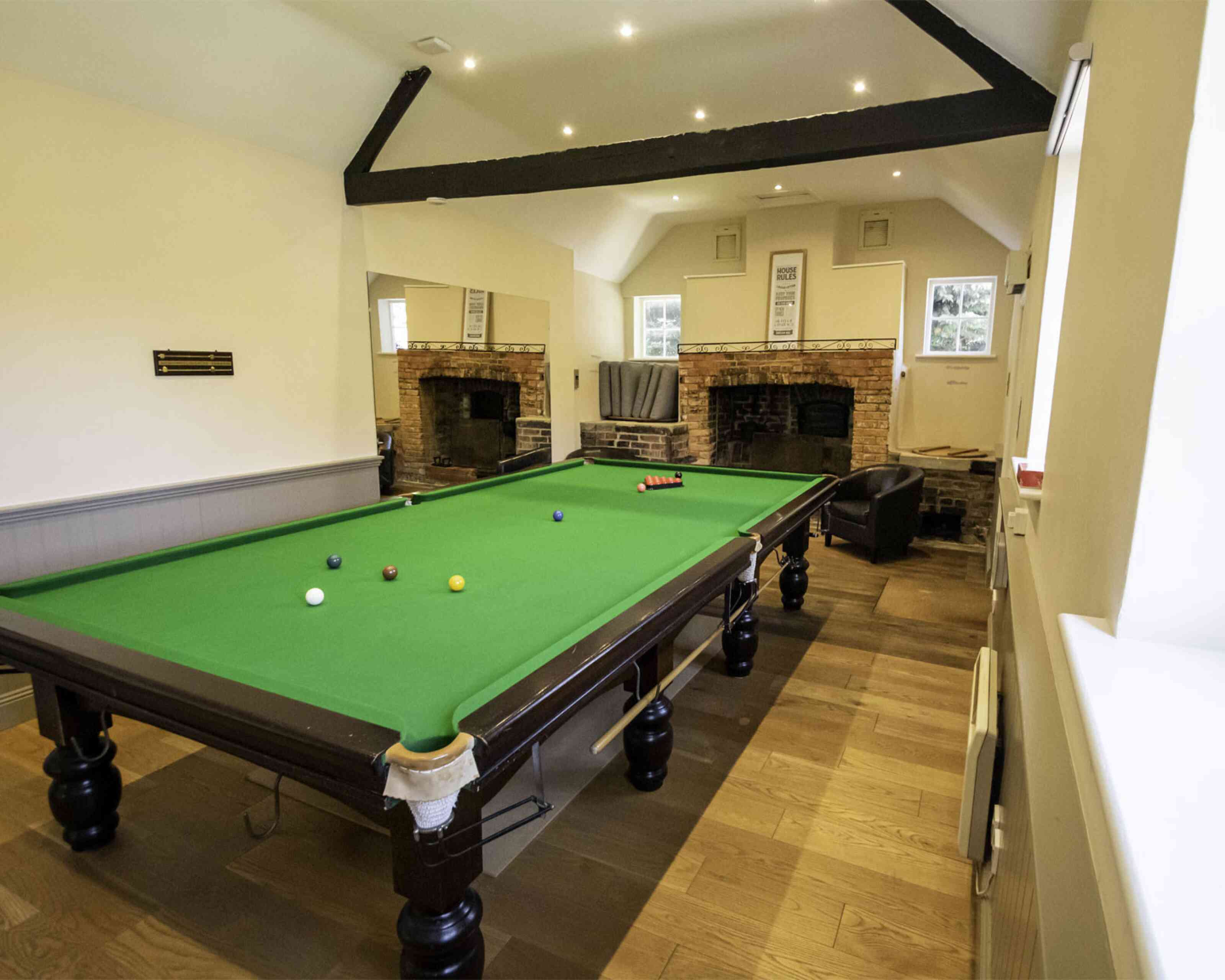 A bright room featuring a green billiards table with balls scattered across it. The walls are light-colored with wooden flooring, and there are two large mirrors reflecting the space. In the background, there are two brick fireplaces and a sofa.