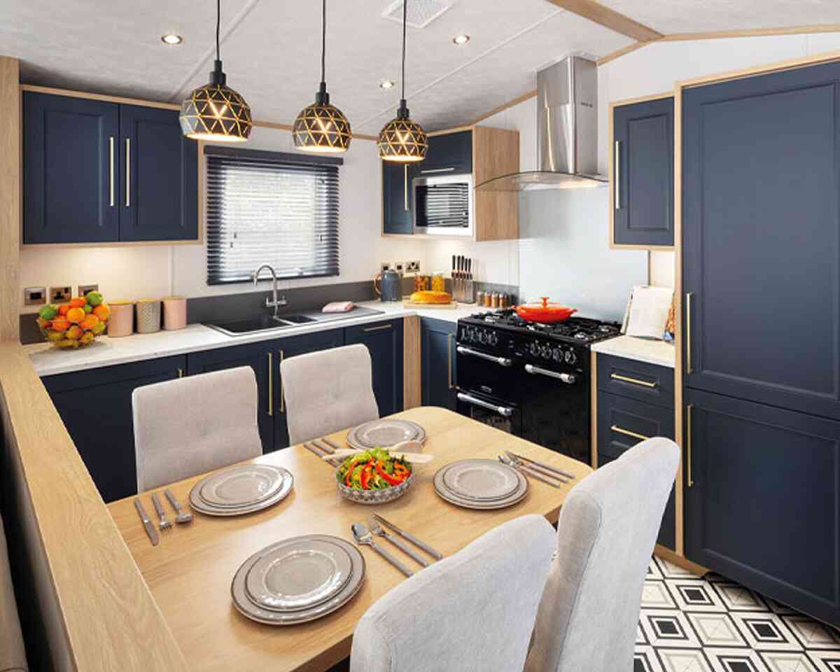 A modern kitchen featuring navy blue cabinets, wooden accents, and a central dining table set for four. Pendant lights hang above the table, and a black range cooker is visible in the background. A colorful salad is placed on the dining table.