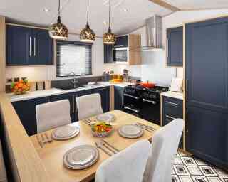 A modern kitchen featuring navy blue cabinets, wooden accents, and a central dining table set for four. Pendant lights hang above the table, and a black range cooker is visible in the background. A colorful salad is placed on the dining table.