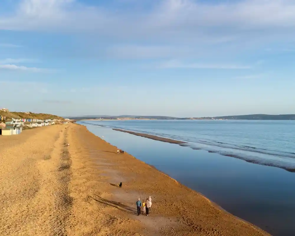 A serene beach at low tide, with soft golden sand and gentle waves. Two people walk along the shoreline, while colorful beach huts line the sand in the distance under a clear blue sky.