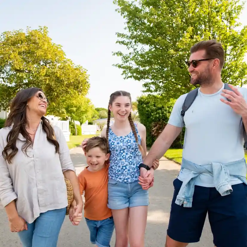 A family of four enjoys a sunny day outdoors, walking along a pathway. The mother and father are smiling and holding hands with their two children, a boy and a girl. The boy, in an orange shirt, playfully runs ahead, while the girl, wearing a blue top, walks beside the mother. Lush greenery and holiday homes are visible in the background.