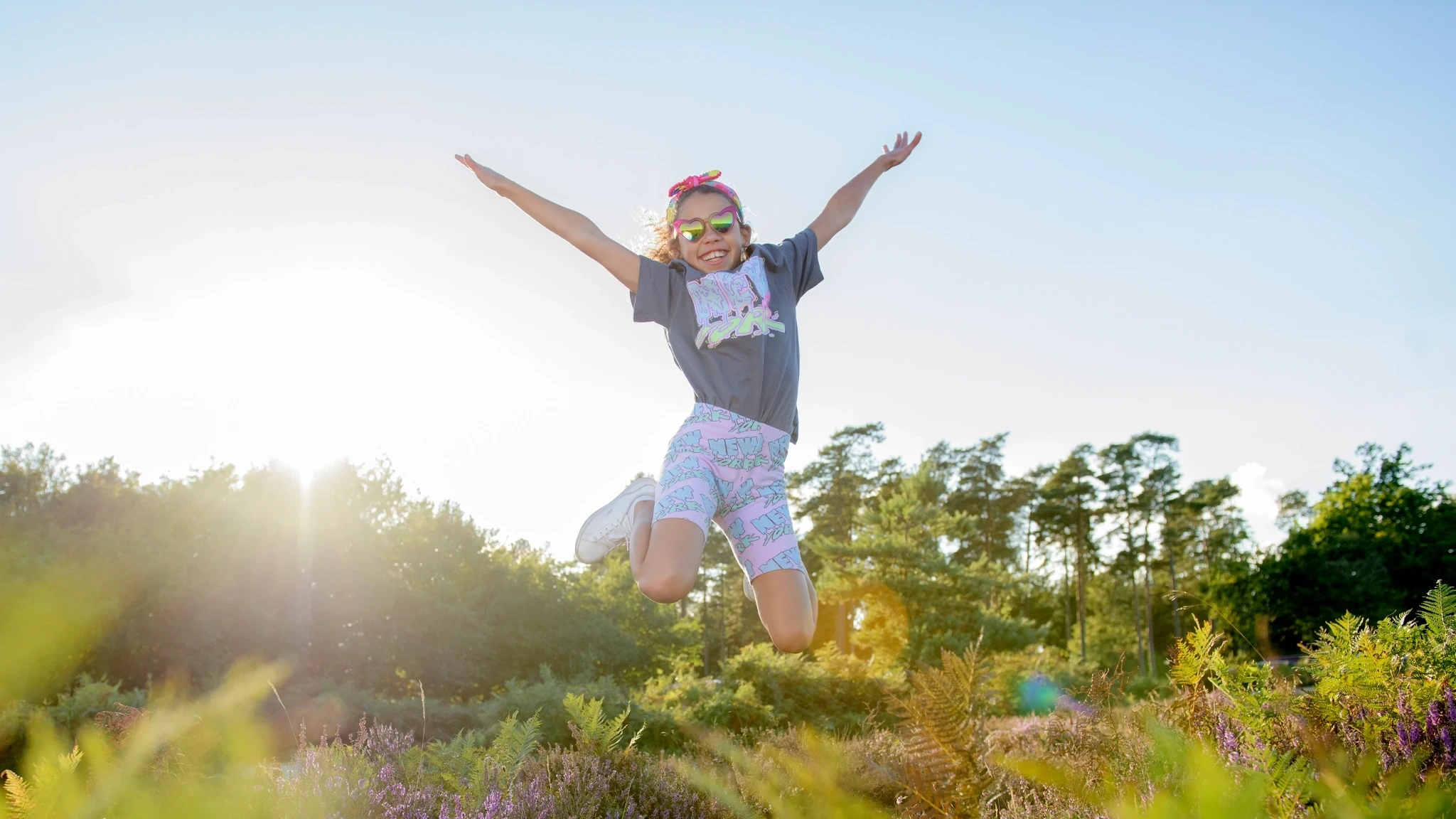 A young girl jumps joyfully in a sunny outdoor setting, wearing a grey t-shirt, colourful shorts, and sunglasses. The background features trees and colourful plants, with sunlight creating a bright, cheerful atmosphere.