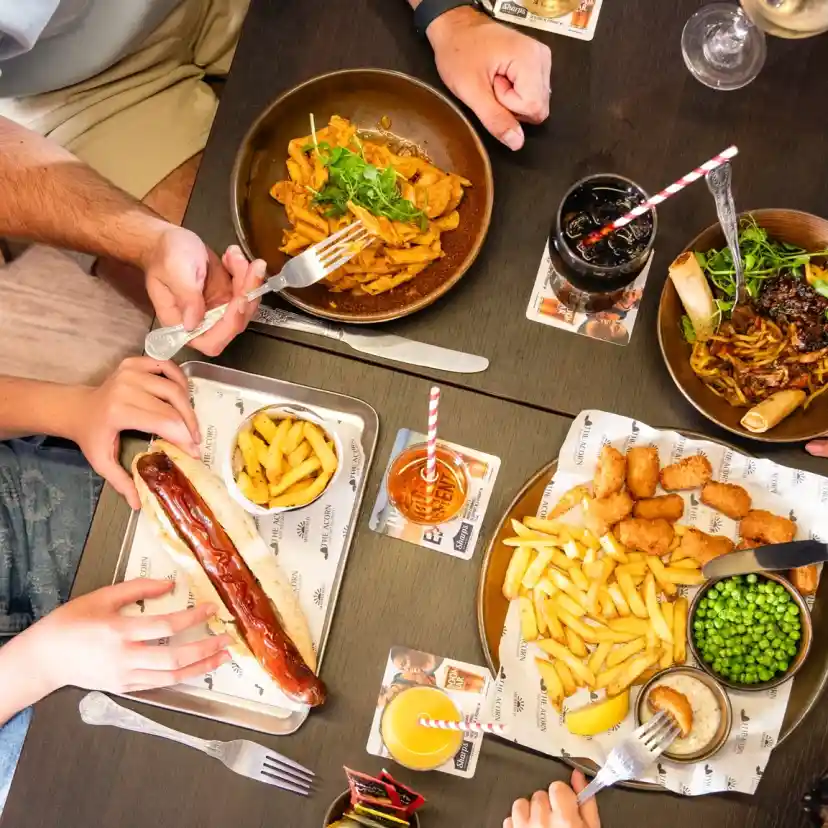 A table set with various dishes, including a large sausage, fries, breaded chicken, green peas, and noodles. Several hands reach for the food, showcasing a casual dining experience.