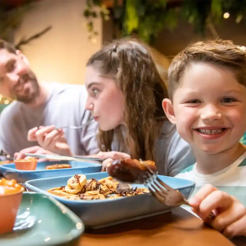 A group of people enjoying a meal at a restaurant, with a smiling young boy in the foreground holding a fork. Others are engaged in conversation while eating off various plates. The ambiance features greenery and warm lighting.