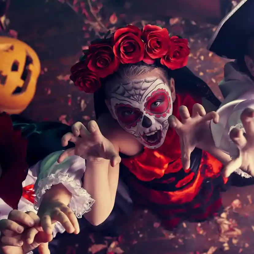 Three children dressed in Halloween costumes reach out playfully. One is a vampire with a cape, another is adorned with a face painted like a sugar skull and red roses, and the third wears a pirate outfit. A carved pumpkin is visible in the background, adding to the festive atmosphere.