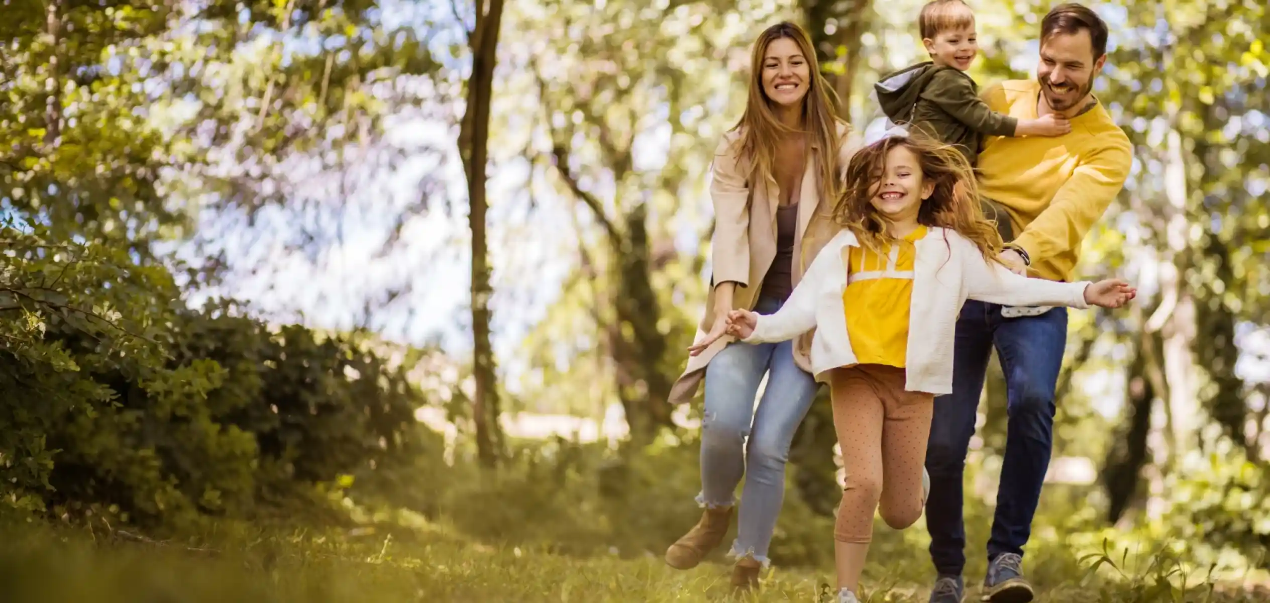 A joyful family runs through a sunlit park, with the mother holding her son on her back while the daughter leads the way, all laughing and enjoying their time together. Lush greenery surrounds them.