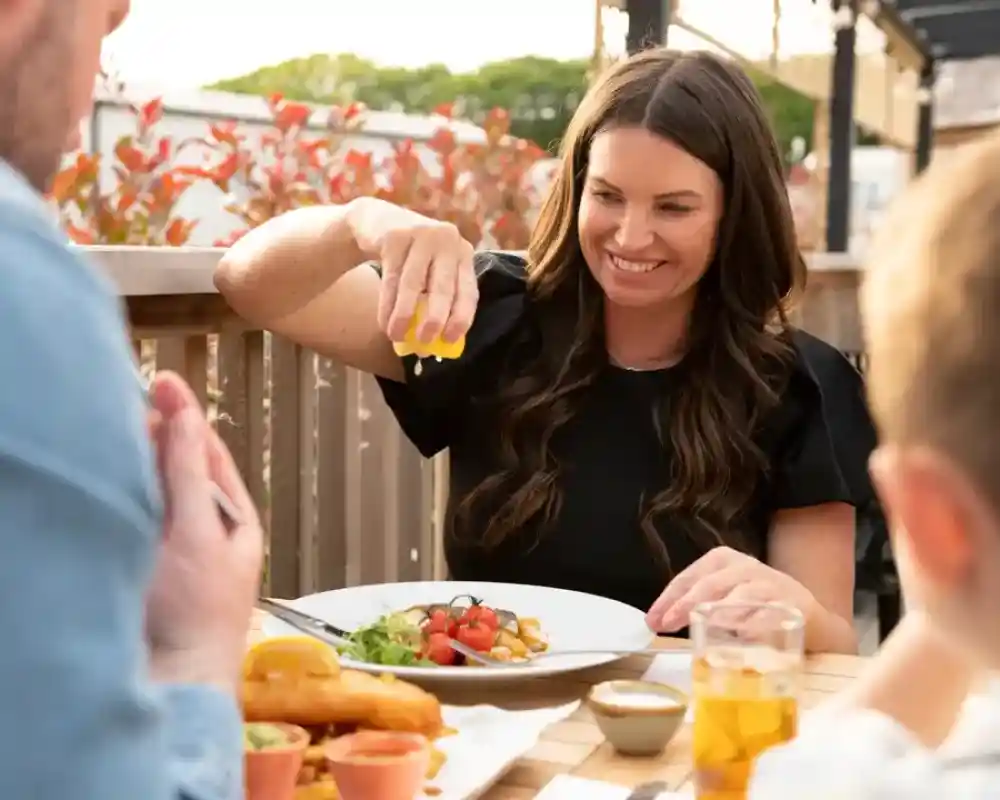 A woman with long, dark hair is squeezing lemon onto her plate while smiling at a casual outdoor dining setting. A child can be seen in the foreground, and a man is in profile, focusing on the meal. Plates of food and drinks are on the table, surrounded by greenery.