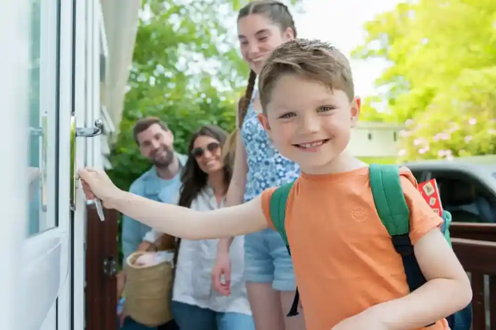A young boy with a backpack smiles as he opens a door, while a group of three adults stands behind him, all looking cheerful. Bright greenery surrounds the scene, suggesting a sunny day.