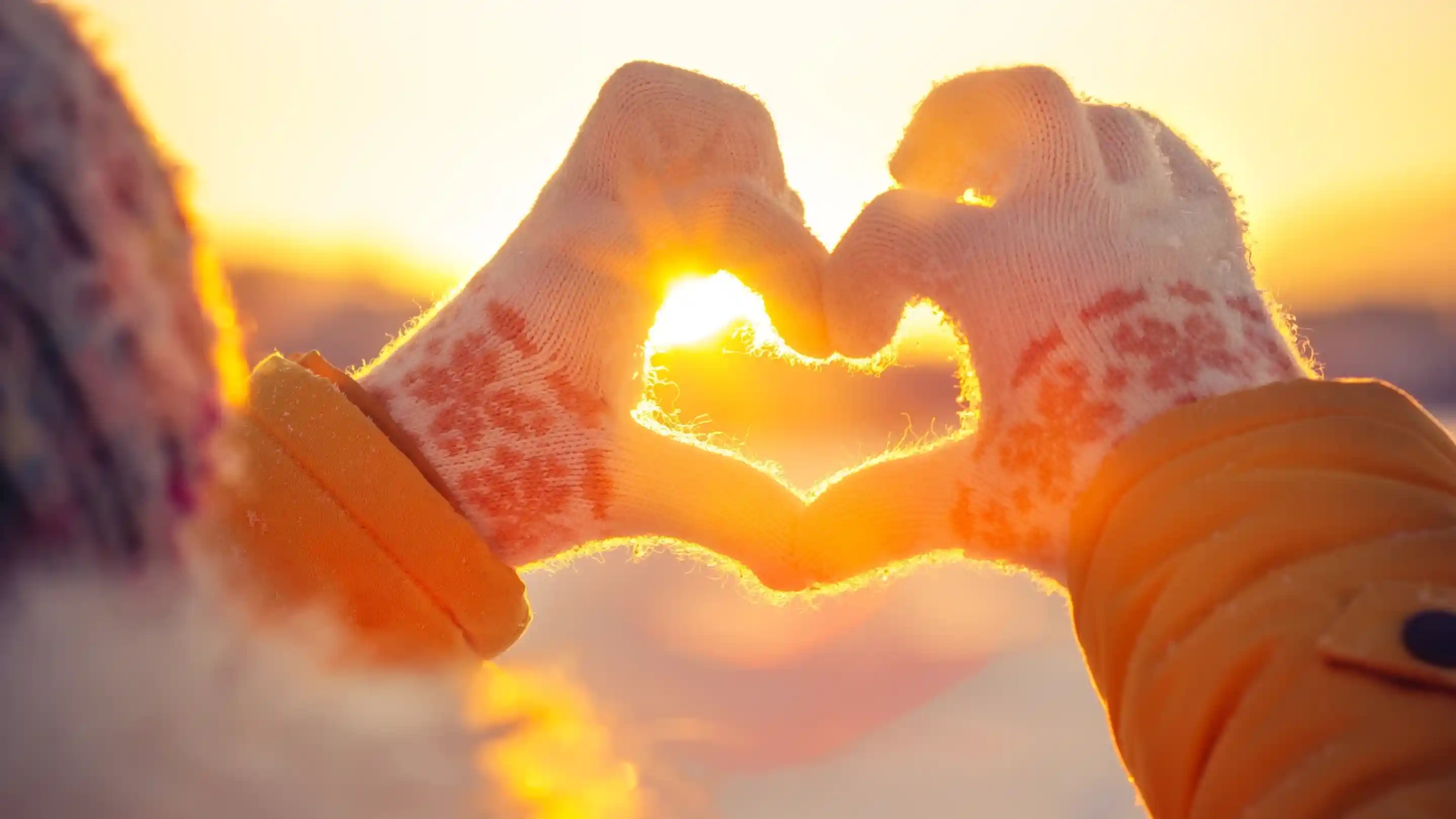 Hands wearing knitted gloves form a heart shape against a bright sunset. The warm glow of the sun shines through the heart, creating a cozy, romantic atmosphere.