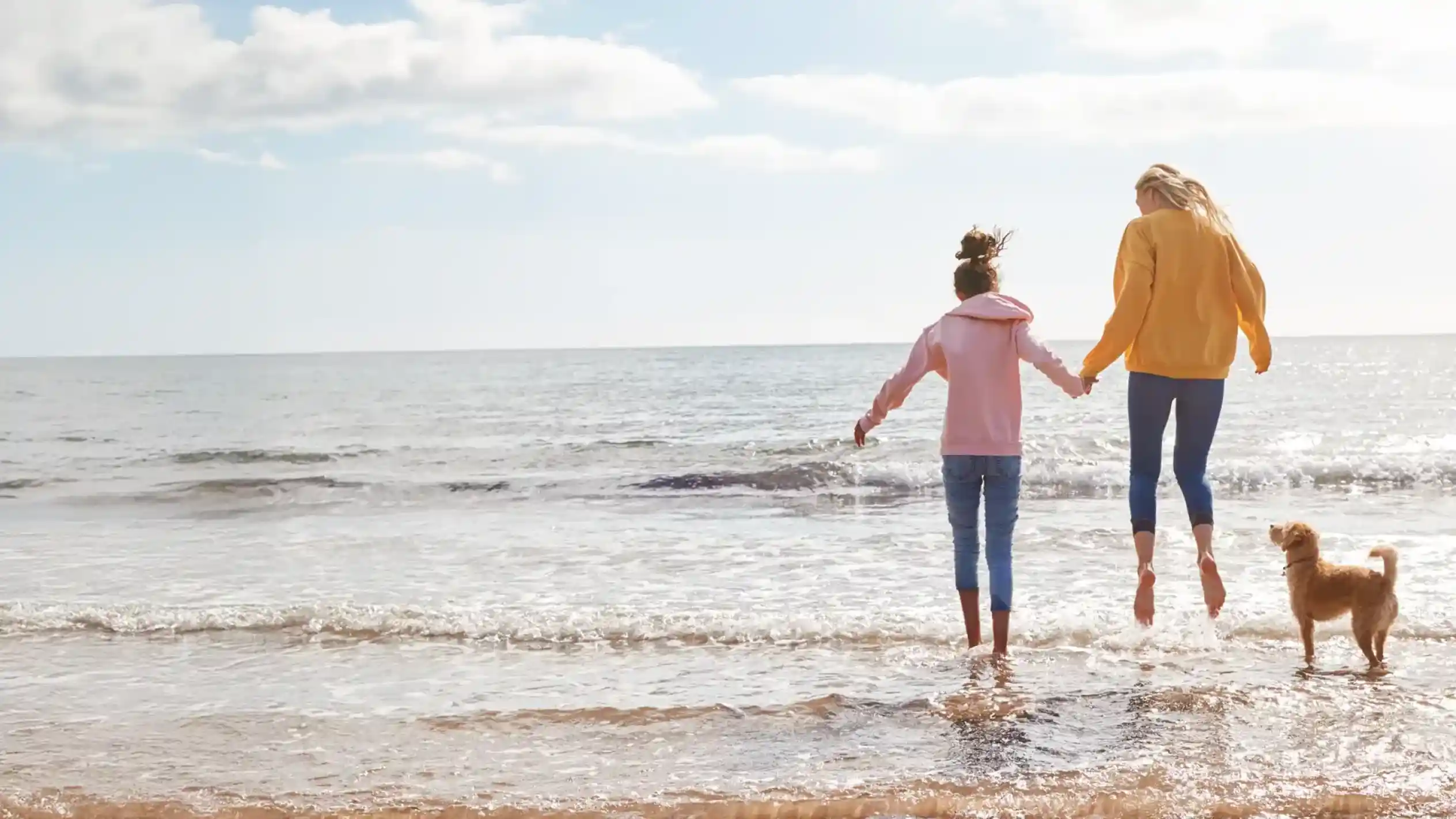 Two women hold hands and jump into the ocean water, with waves splashing around their feet. A small dog stands nearby on the beach, enjoying the scene under a clear sky with fluffy clouds.