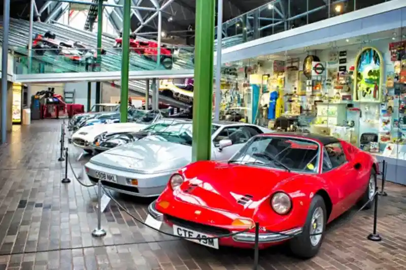 A collection of classic cars on display in a museum setting, featuring a prominent red sports car in the foreground and several other vehicles lined up behind it. The backdrop includes colourful displays and exhibits.
