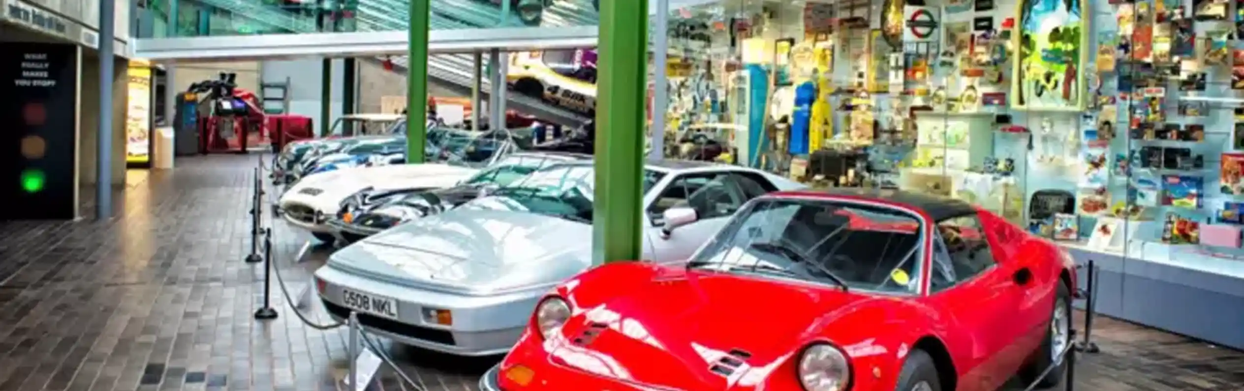 A collection of classic cars on display in a museum setting, featuring a prominent red sports car in the foreground and several other vehicles lined up behind it. The backdrop includes colourful displays and exhibits.