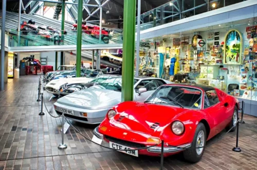 A collection of classic cars on display in a museum setting, featuring a prominent red sports car in the foreground and several other vehicles lined up behind it. The backdrop includes colourful displays and exhibits.