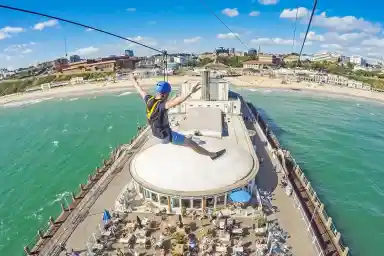 A person enjoys a zip line ride over a beachside pier, arms outstretched, with a vibrant blue sky and green ocean visible below. The pier features outdoor seating and people enjoying the sun.