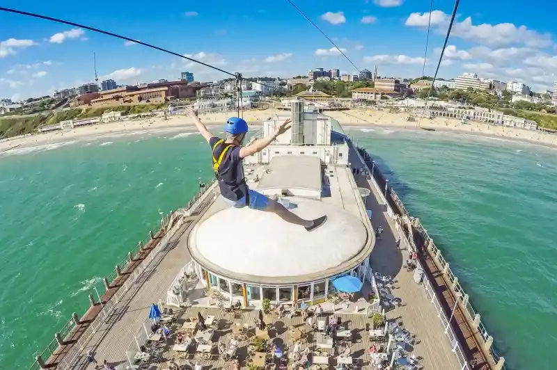 A person enjoys a zip line ride over a beachside pier, arms outstretched, with a vibrant blue sky and green ocean visible below. The pier features outdoor seating and people enjoying the sun.