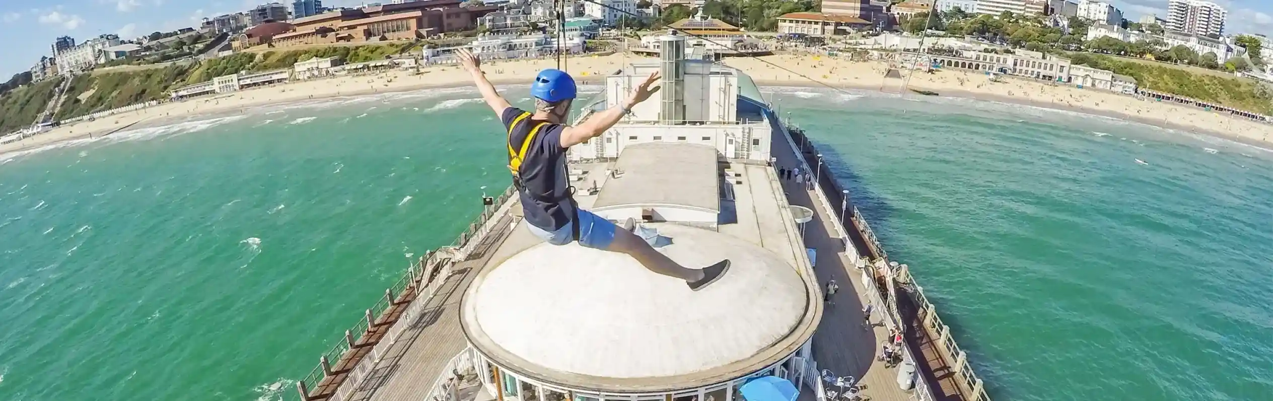 A person enjoys a zip line ride over a beachside pier, arms outstretched, with a vibrant blue sky and green ocean visible below. The pier features outdoor seating and people enjoying the sun.