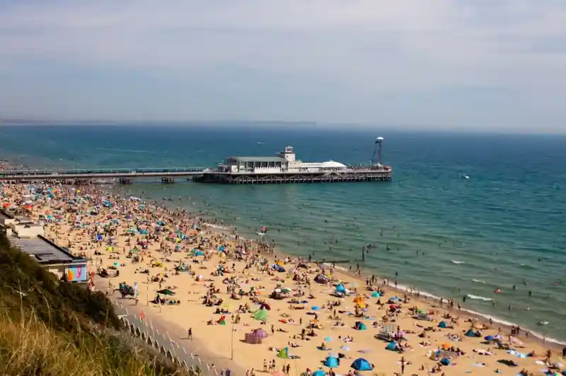 A crowded beach filled with colourful umbrellas and sunbathers. A pier extends over the water in the background, with calm waves and a clear blue sky above.