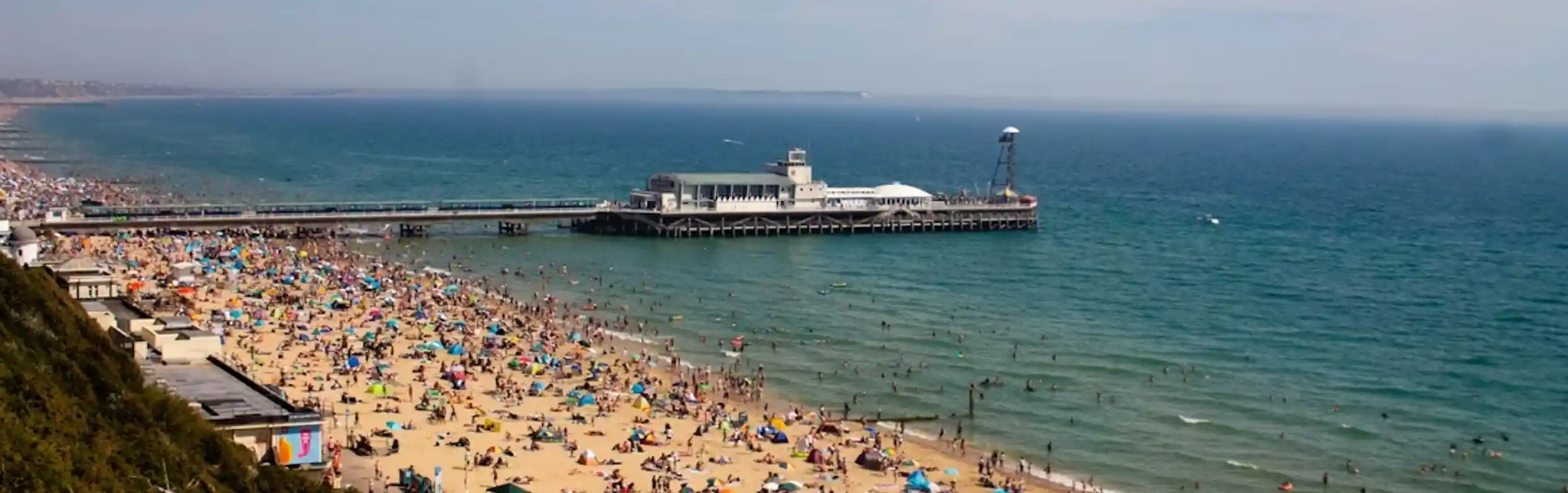 A crowded beach filled with colourful umbrellas and sunbathers. A pier extends over the water in the background, with calm waves and a clear blue sky above.