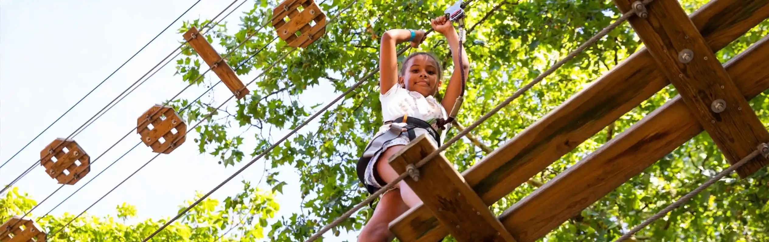 A child is confidently navigating through a ropes course, suspended high among trees, while wearing a safety harness. Sunlight filters through the leaves, illuminating the scene.