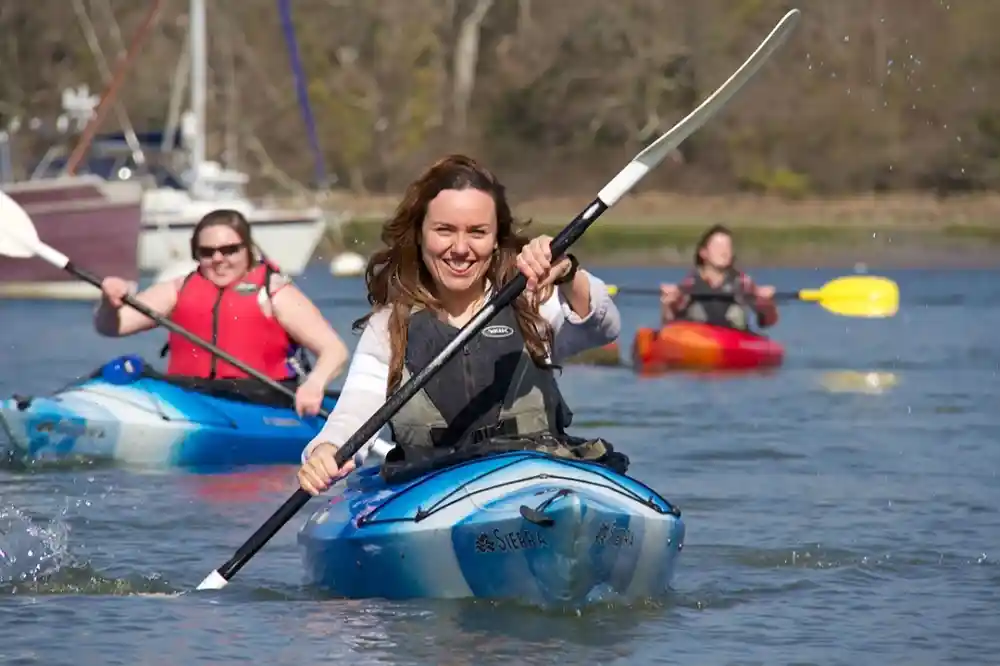 A woman joyfully paddles a kayak on a calm waterway, smiling at the camera. Two other kayakers are visible in the background, enjoying the sunny day. Boats are docked along the shore.