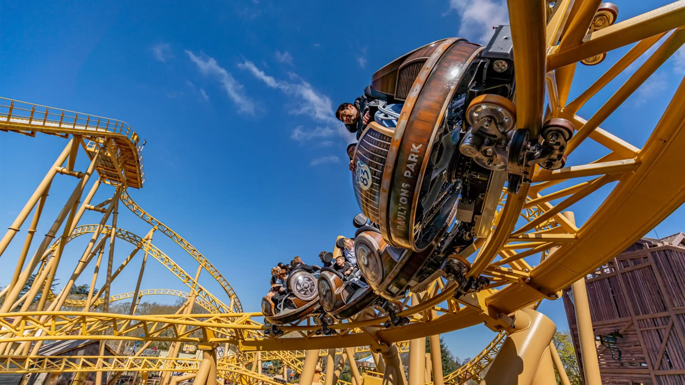 A roller coaster with vintage-style cars zooms through a loop under a clear blue sky. Riders are enjoying the thrill, with some raising their arms in excitement. The track is a bright yellow, contrasting with the surrounding landscape.