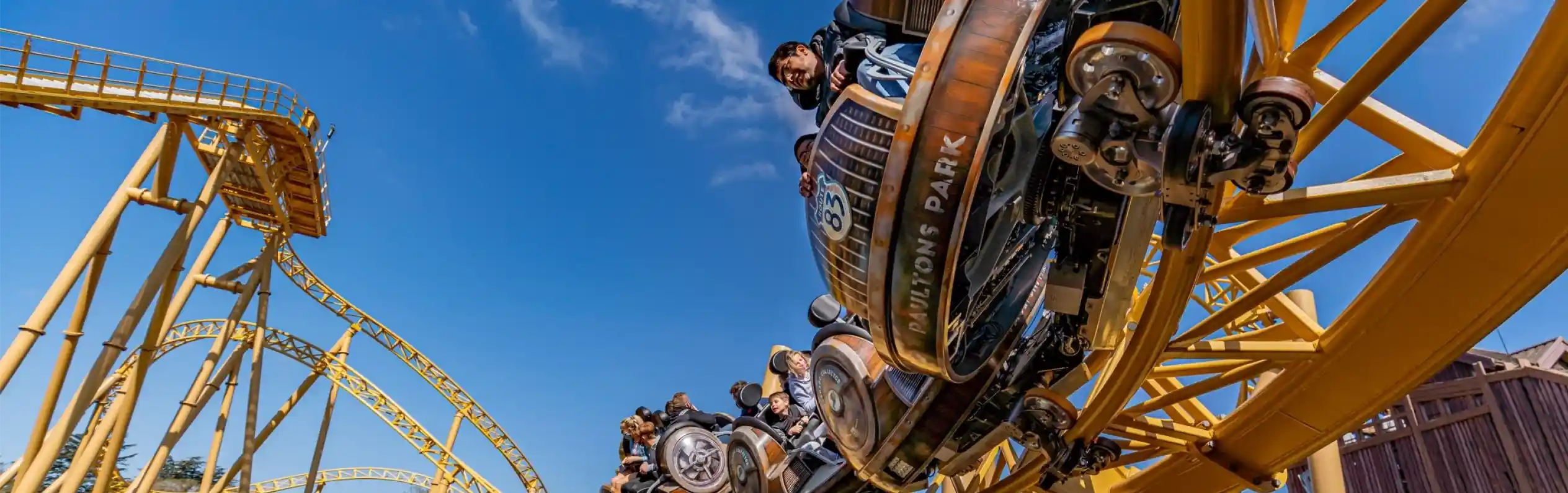 A roller coaster with vintage-style cars zooms through a loop under a clear blue sky. Riders are enjoying the thrill, with some raising their arms in excitement. The track is a bright yellow, contrasting with the surrounding landscape.