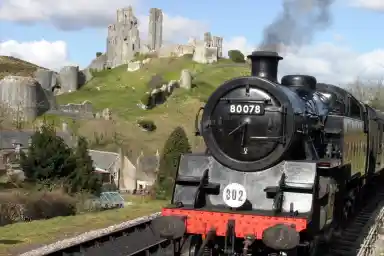 A steam train approaches a historic castle on a hill, surrounded by lush greenery and a partly cloudy sky. The train emits a plume of smoke, creating a nostalgic atmosphere.