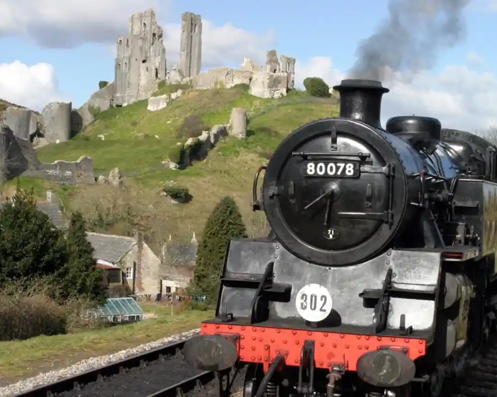 A steam train approaches a historic castle on a hill, surrounded by lush greenery and a partly cloudy sky. The train emits a plume of smoke, creating a nostalgic atmosphere.