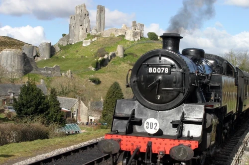 A steam train approaches a historic castle on a hill, surrounded by lush greenery and a partly cloudy sky. The train emits a plume of smoke, creating a nostalgic atmosphere.