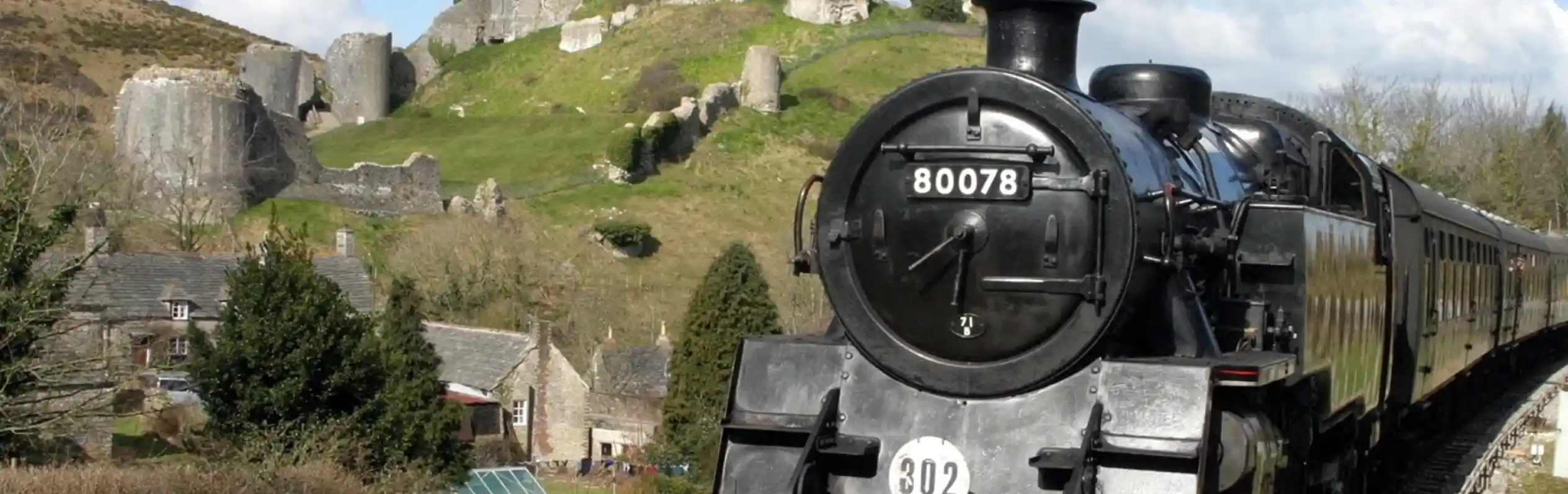 A steam train approaches a historic castle on a hill, surrounded by lush greenery and a partly cloudy sky. The train emits a plume of smoke, creating a nostalgic atmosphere.