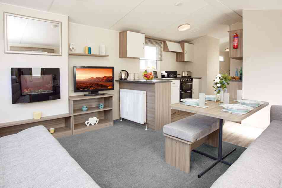 The image shows the interior of a bright, modern caravan or mobile home, featuring an open-plan lounge, dining, and kitchen area. In the lounge section, there’s a wall-mounted electric fireplace with a reflective glass surface, a flat-screen TV displaying a vivid desert landscape, and light wood shelving holding decorative items, including teal candle holders and a white heart-shaped ornament. The dining area has a wooden table set with four plates, glasses, placemats, and a vase of fresh flowers, accompanied by cushioned benches upholstered in light grey fabric. The kitchen, located behind the dining space, has light wood and white cabinetry, a countertop with a kettle and a bowl of fruit, and a black freestanding oven with a gas hob. A fire extinguisher is mounted on the wall in the background. The space is decorated in neutral tones with light grey carpet in the lounge and wood-effect flooring in the kitchen and dining areas.
