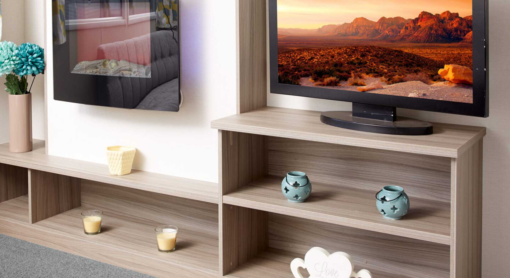 The image shows a close-up of a modern caravan or mobile home entertainment area. On the left, there is a sleek wall-mounted electric fireplace with a reflective glass surface, showing the reflection of a sofa and patterned curtains. Below it is a long wooden shelf holding a vase with blue artificial flowers, a small yellow candle holder, and two tealight candles. On the right, a flat-screen TV displays a vivid landscape of rocky mountains under an orange sky. The TV sits on a light wood shelving unit that contains two turquoise decorative candle holders on the middle shelf and a white heart-shaped ornament with the words “Love the life you live” on the bottom shelf. The floor is carpeted in soft grey.