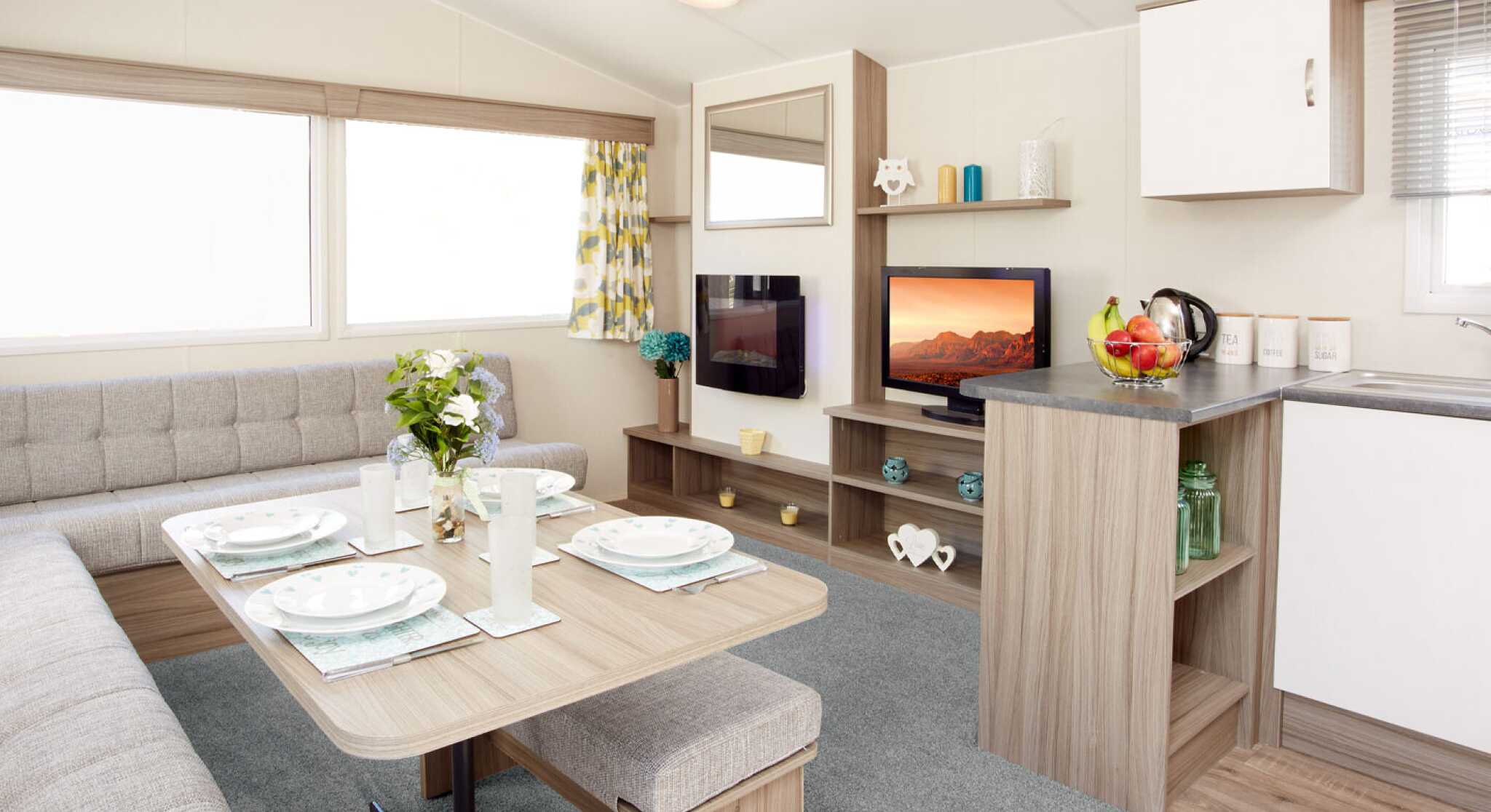 The image shows the interior of a modern caravan or mobile home. It features an L-shaped light-grey upholstered seating area surrounding a wooden dining table set with four plates, glasses, and placemats. A vase of fresh flowers sits in the centre of the table. To the right, there’s a compact kitchen area with a grey countertop holding a fruit bowl, a kettle, and labelled storage canisters. Behind the counter is a small entertainment area with a flat-screen TV displaying a scenic landscape, shelves with decorative items, and a large window with patterned yellow-and-grey curtains letting in natural light. The floor is a mix of light wood and a soft grey carpet in the lounge area.