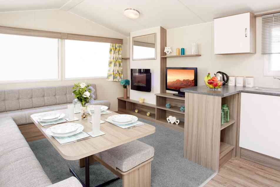The image shows the interior of a modern caravan or mobile home. It features an L-shaped light-grey upholstered seating area surrounding a wooden dining table set with four plates, glasses, and placemats. A vase of fresh flowers sits in the centre of the table. To the right, there’s a compact kitchen area with a grey countertop holding a fruit bowl, a kettle, and labelled storage canisters. Behind the counter is a small entertainment area with a flat-screen TV displaying a scenic landscape, shelves with decorative items, and a large window with patterned yellow-and-grey curtains letting in natural light. The floor is a mix of light wood and a soft grey carpet in the lounge area.