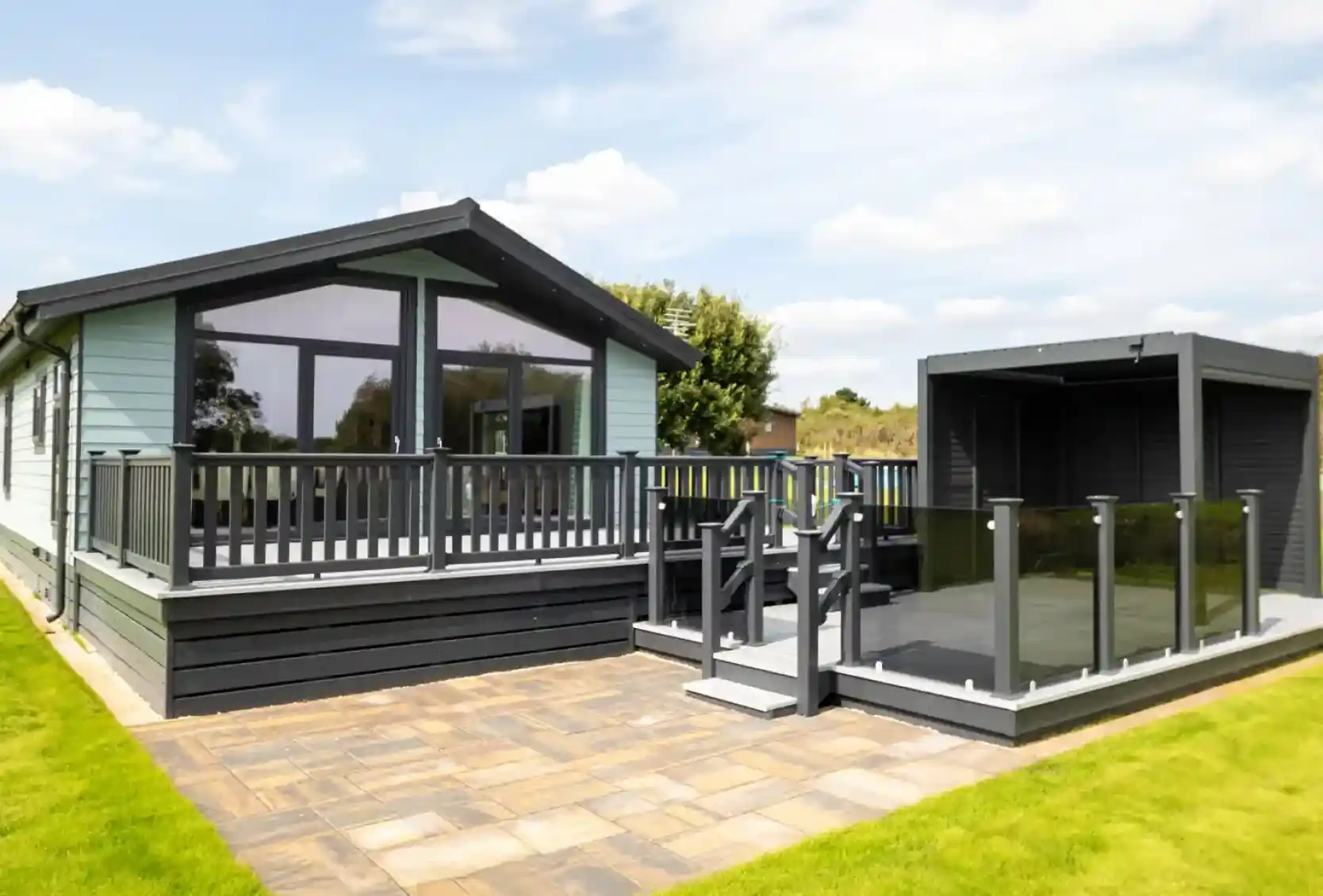 Modern light blue holiday home with a grey wraparound deck and glass balustrades, adjacent to a dark grey outdoor pergola.