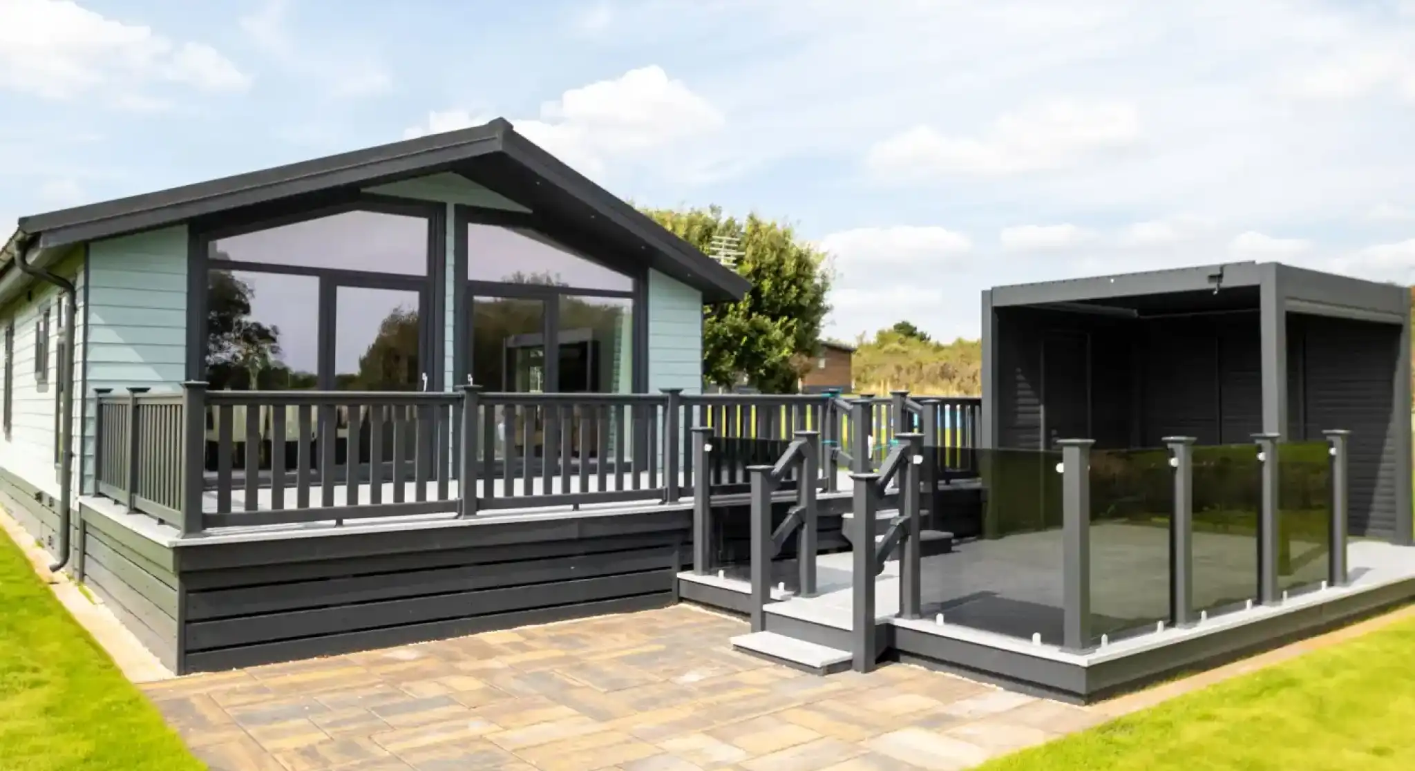 Modern light blue holiday home with a grey wraparound deck and glass balustrades, adjacent to a dark grey outdoor pergola.